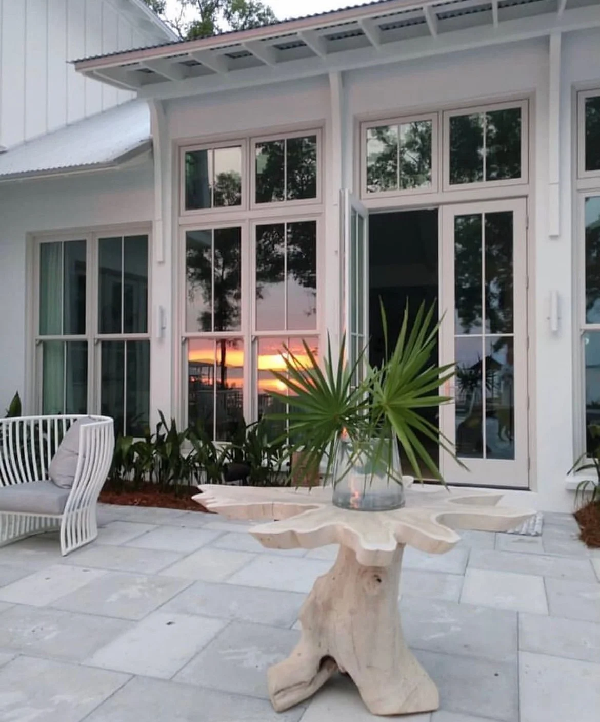 Patio with a wooden table, tropical plant in a glass vase, and outdoor furniture, with a sunset reflecting in large window panes of a modern house.