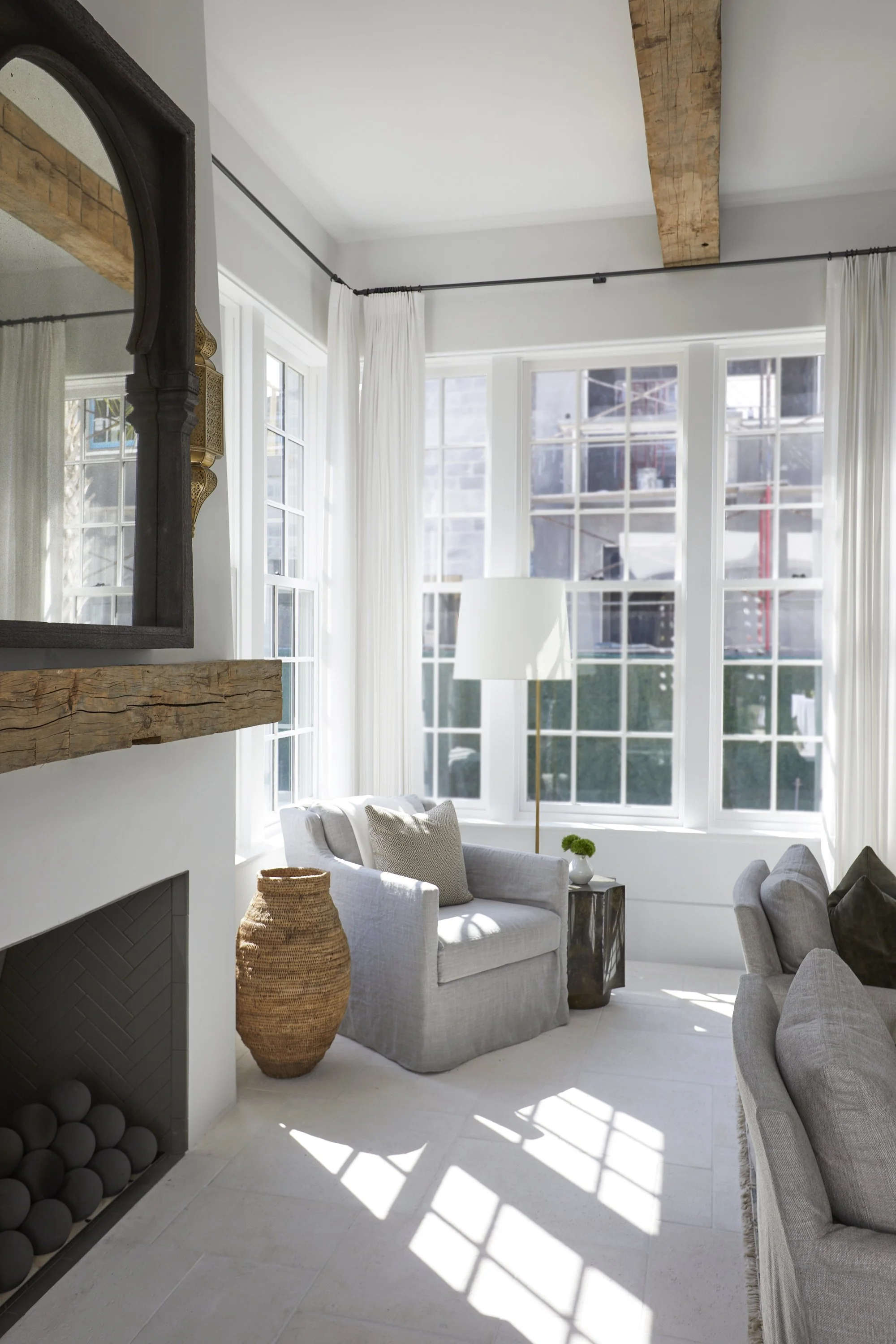 Bright living room with large windows, white curtains, gray armchair with pillow, wicker basket, white lamp, and stone fireplace.
