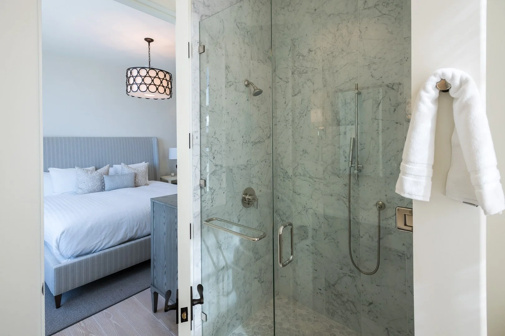 View from a bathroom into a bedroom, showing a walk-in shower with marble tiles, a towel hanging on the door, and a bed with a headboard and pillows in the bedroom.