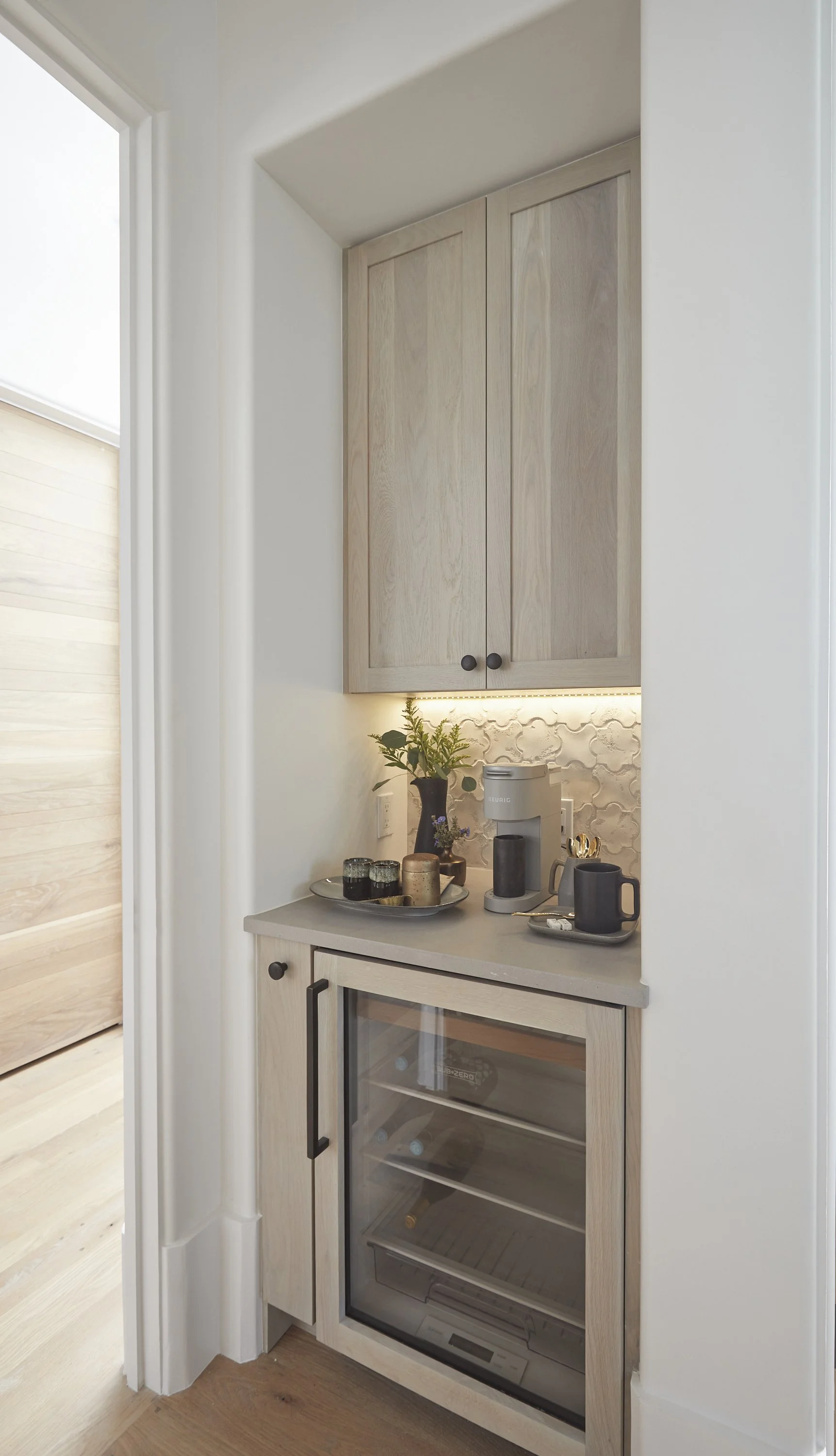 A small kitchen nook with a beige countertop, a toaster, a coffee machine, and cups on a tray. There are wooden cabinets above and a mini fridge below, with a decorative backsplash and a small plant.