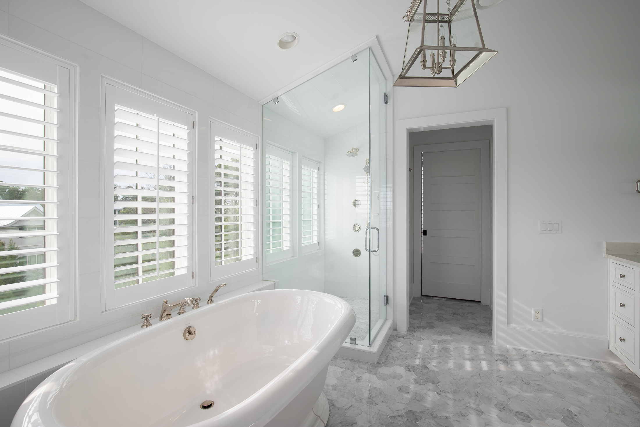 Bright bathroom featuring a white bathtub near multiple windows with white shutters, a glass-enclosed shower, a white door, and a pendant light fixture.