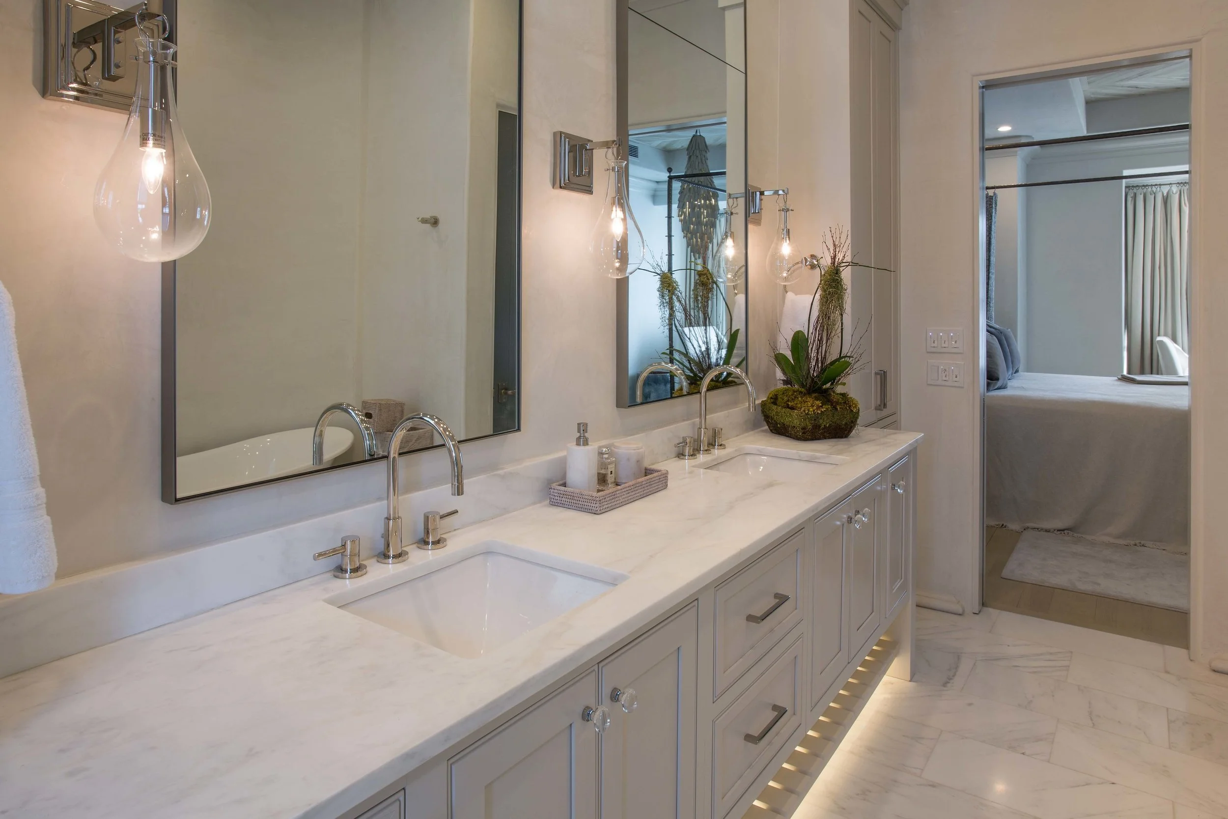 A bathroom with a double vanity with a white marble countertop, two rectangular sinks, and two mirrored medicine cabinets. There are three wall-mounted glass hanging light fixtures above the mirror, and a decorative plant on the counter. The bathroom