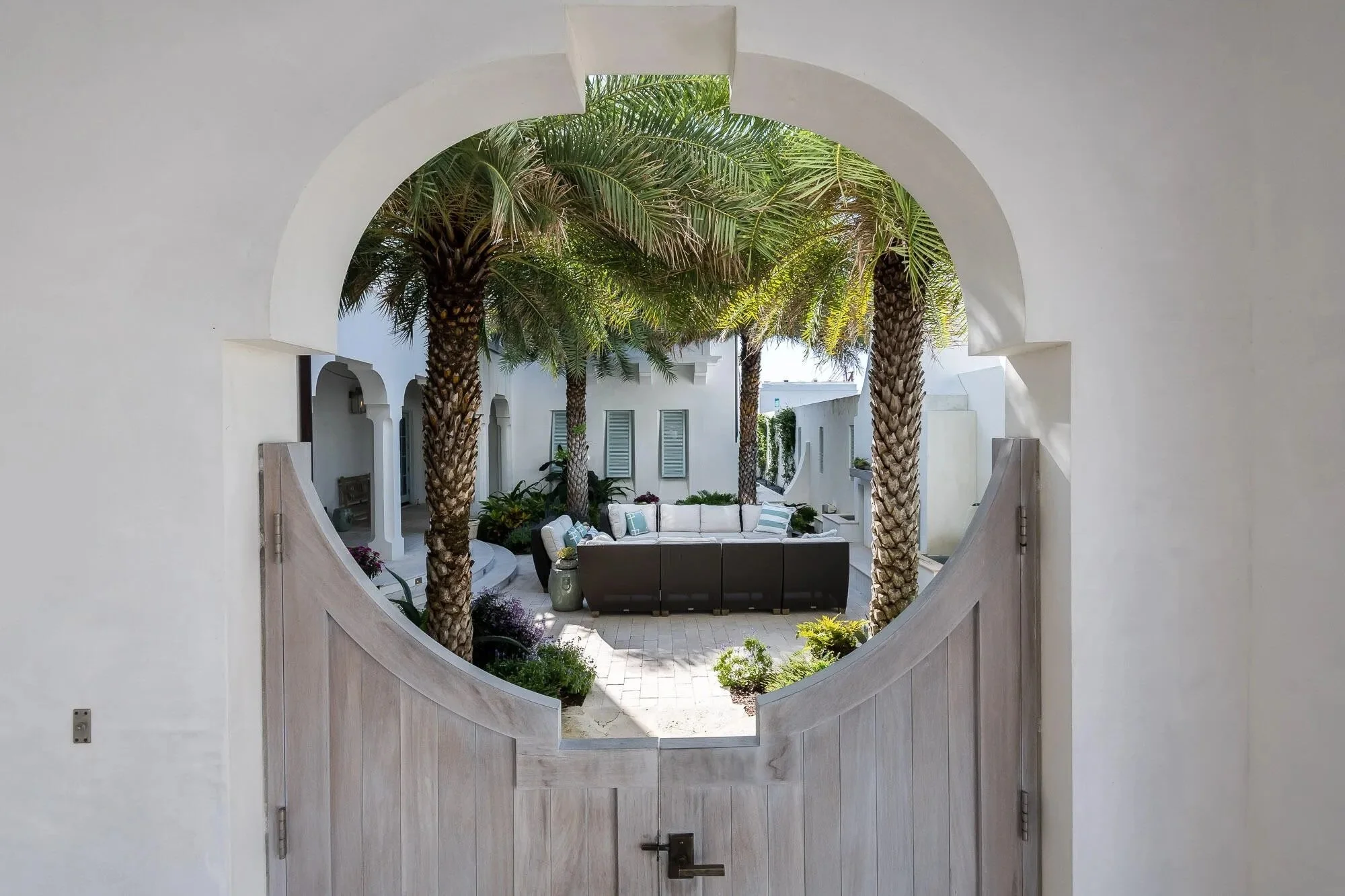 View through a wooden gate opening to an outdoor courtyard with white walls, tall palm trees, outdoor seating, and potted plants.