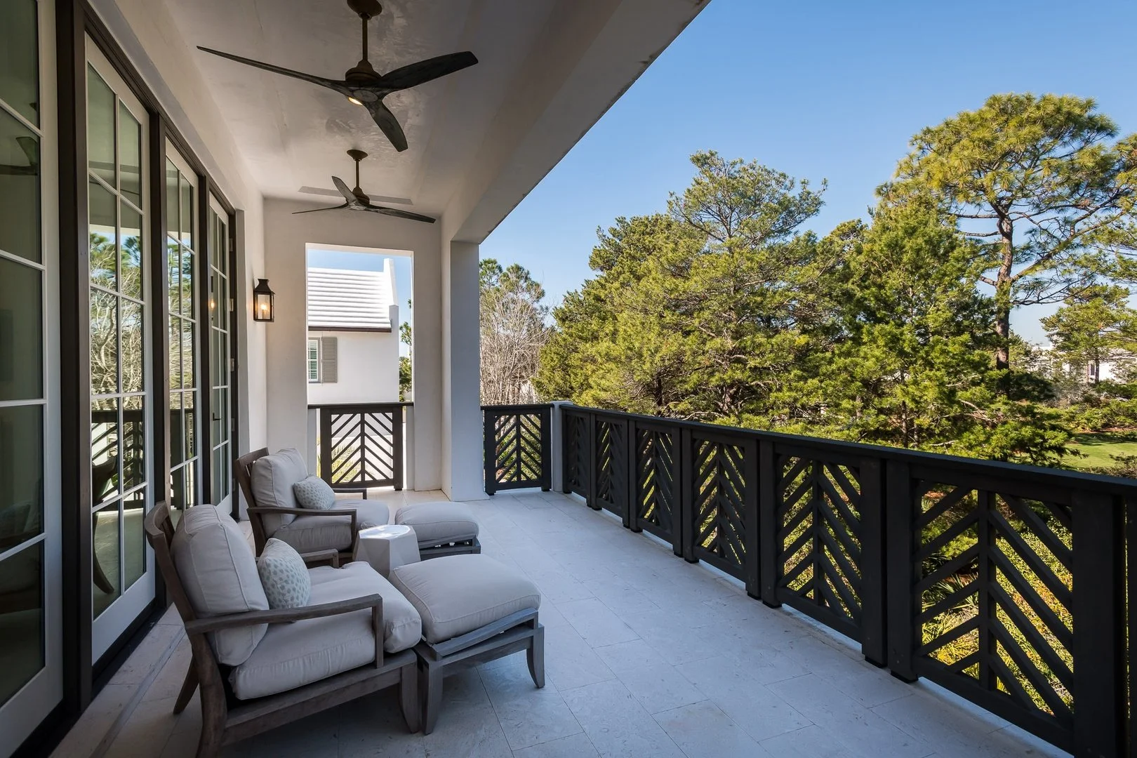Balcony with white cushioned chairs and a small table, black railings, view of trees and blue sky.