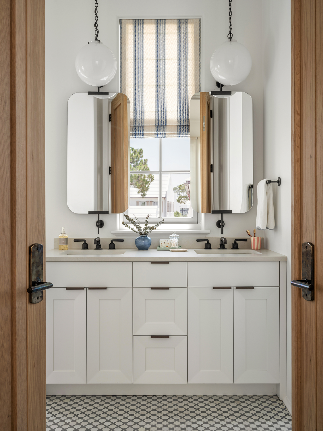 View of a bathroom with two white sinks, black fixtures, two mirrors, hanging globe lights, a window with a striped blind, a blue vase with greenery, and a patterned floor.