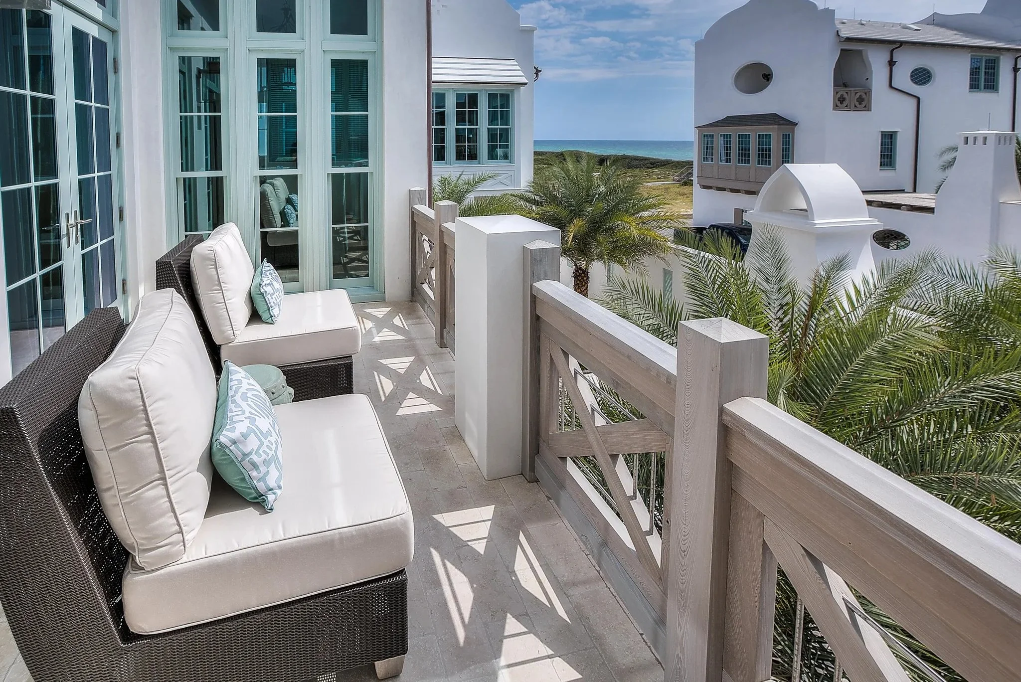 Balcony with white cushioned chairs and pillows overlooking palm trees, neighboring elegant white buildings, and the ocean in the distance.