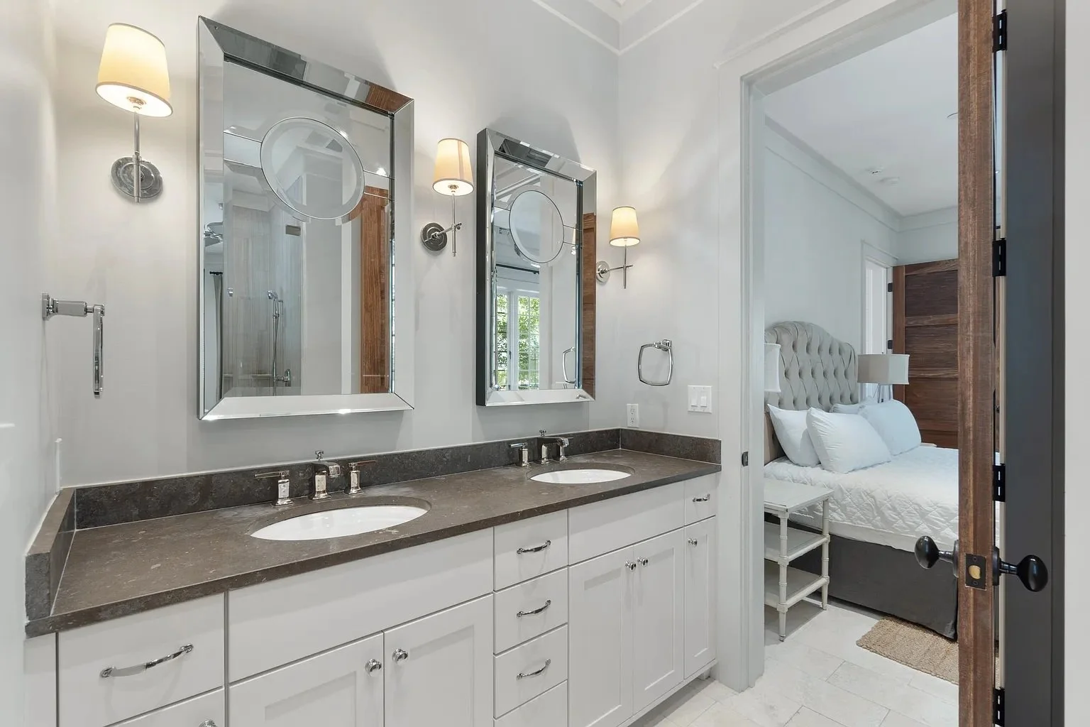 Bathroom vanity with two sinks, mirrors, wall-mounted lights, and a view into a bedroom with a bed and nightstand.