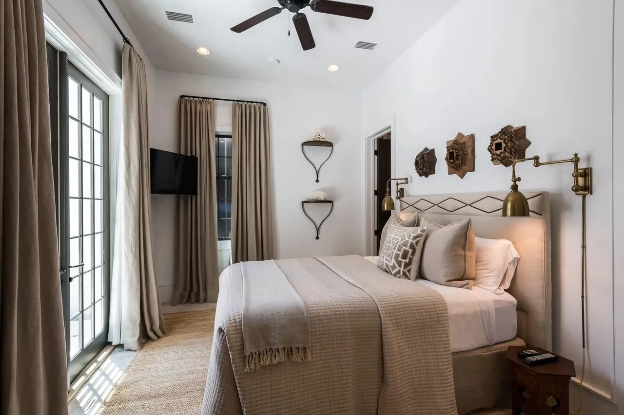 Bedroom with a large bed, beige headboard, beige and white bedding, pillows, wall-mounted reading lamps, decorative wall art, a small side table, and a TV, with beige curtains and glass door leading outside.