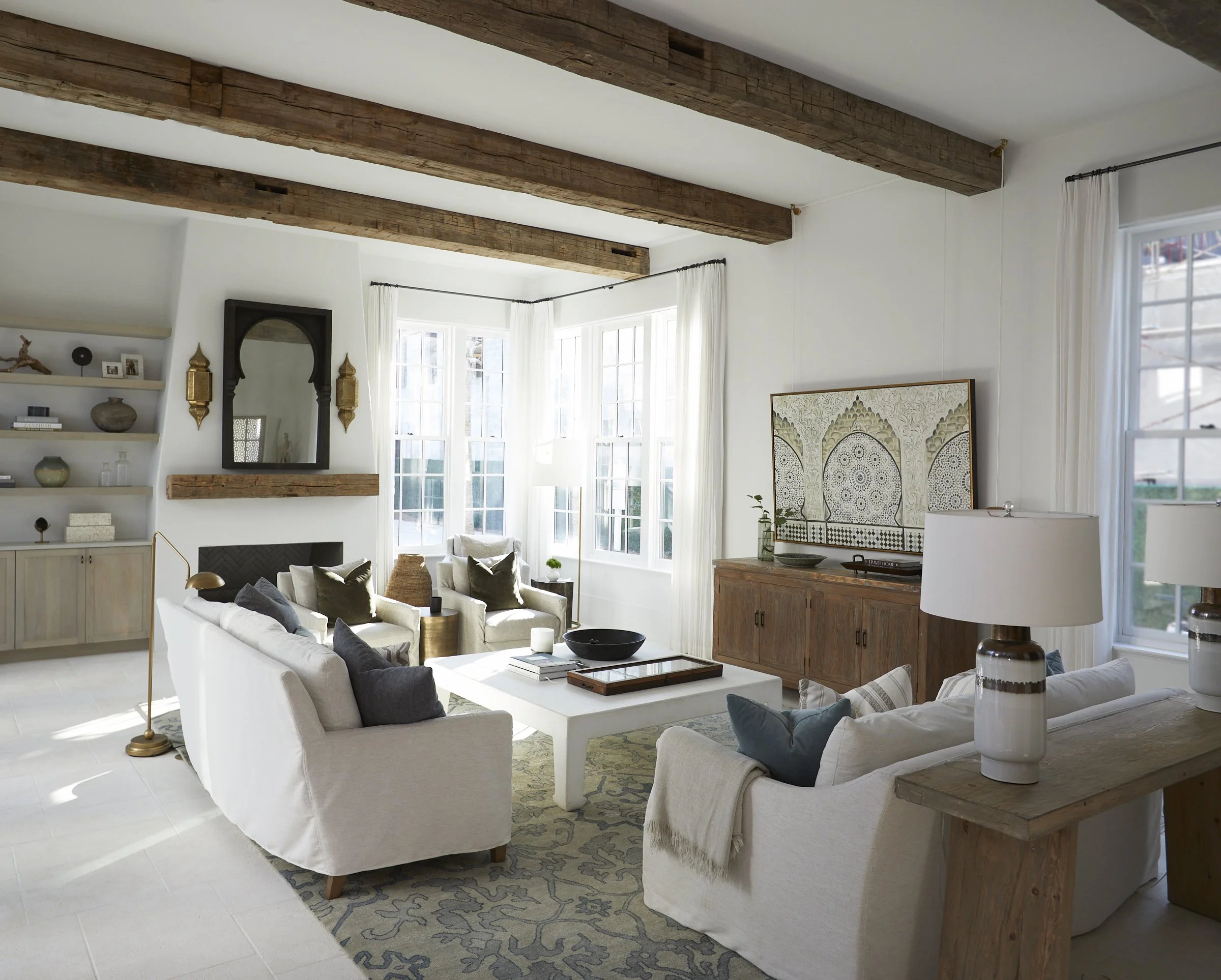 Bright living room with white sofas, wooden ceiling beams, and large windows, decorated in a neutral color palette with pillows, lamps, and artwork.