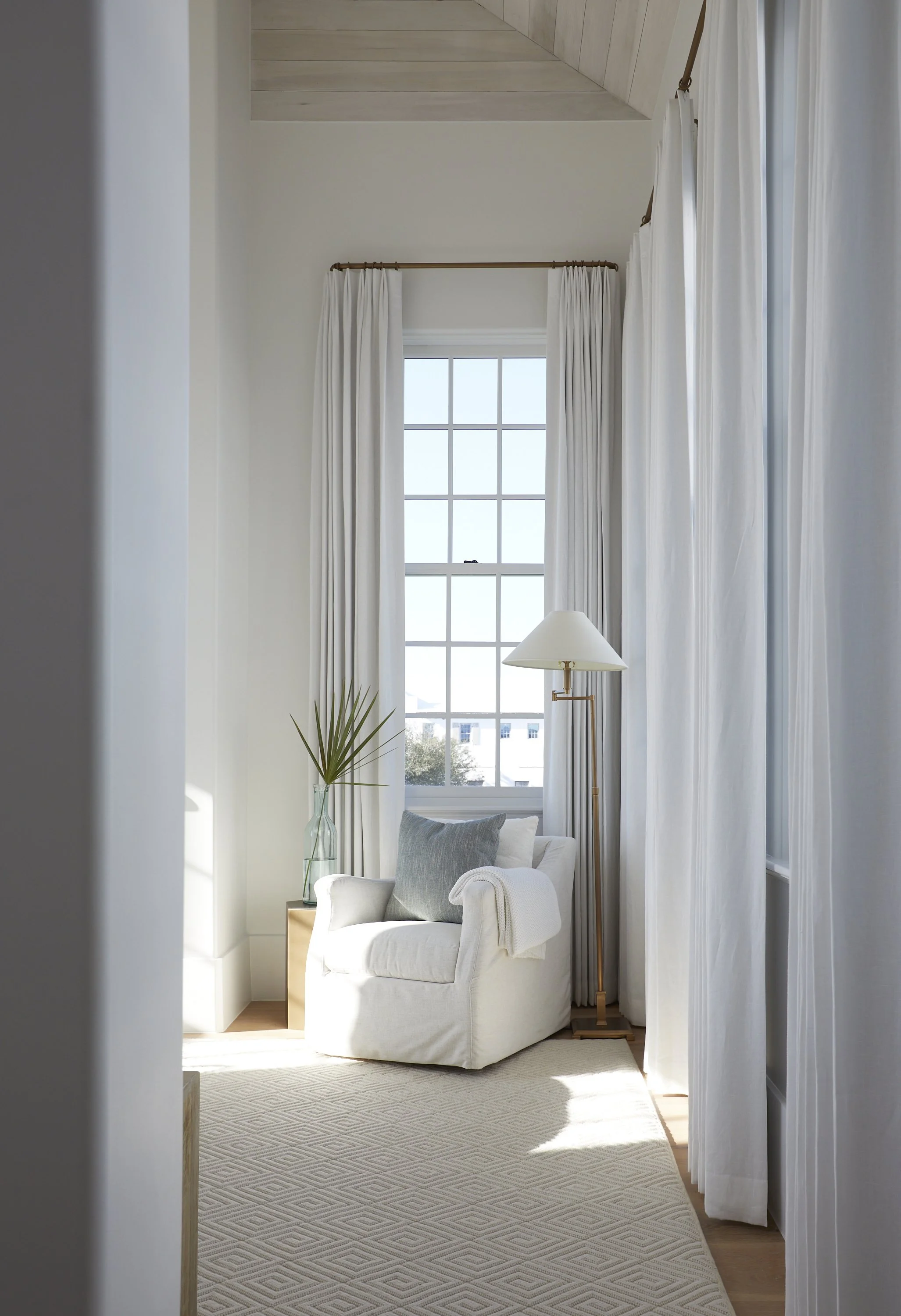 A cozy corner of a bright living room with a white armchair, a gray pillow, a white blanket, a tall window with white curtains, a floor lamp, a small side table with a glass vase and plant, and a light-colored rug.