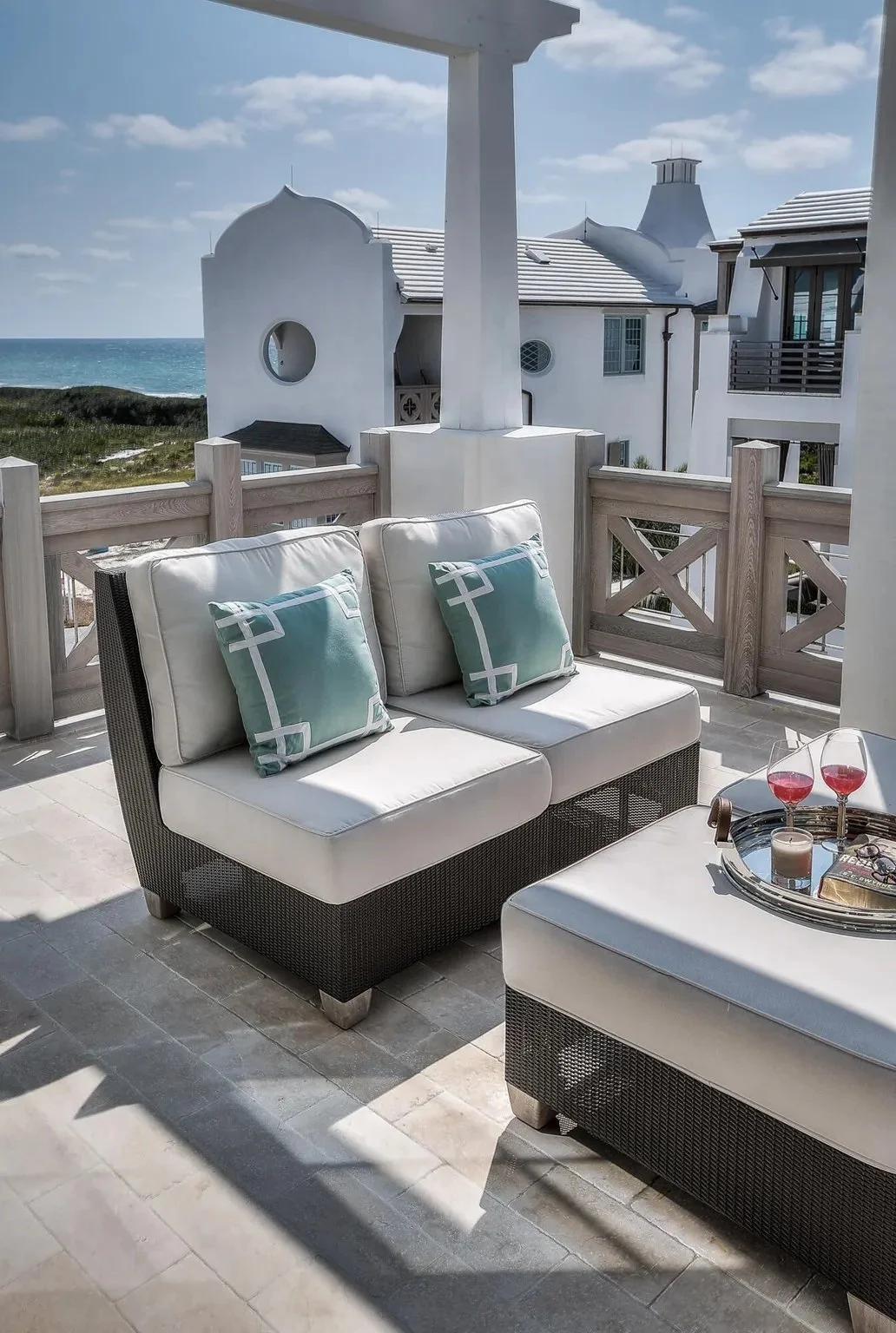 A balcony with two cushioned wicker sofas, each with two teal pillows, overlooking a coastal scene with white buildings and the ocean in the background.