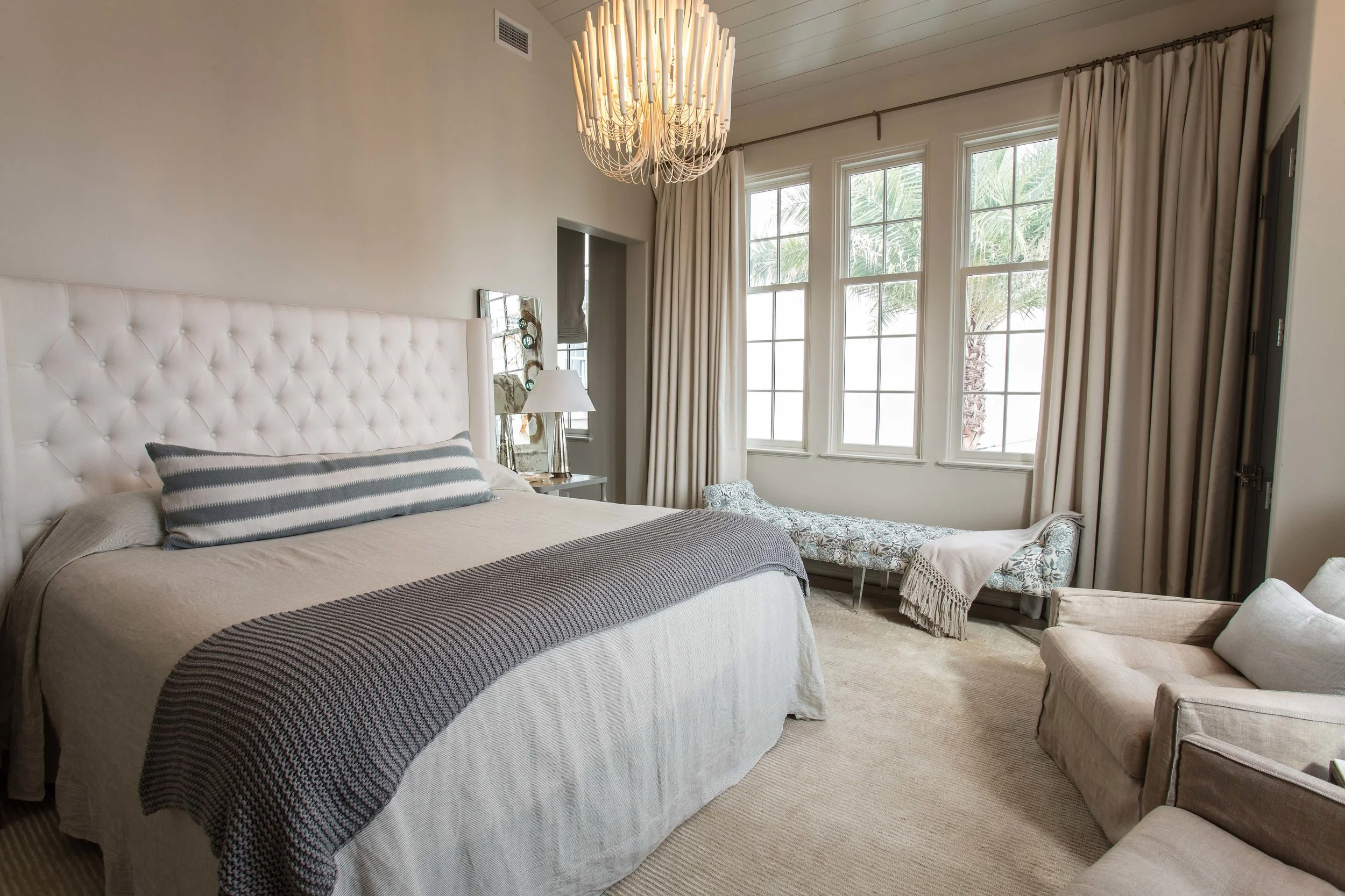 A bedroom with a large white upholstered bed, gray and white striped pillows, beige bedding with a textured gray blanket, a beige armchair, a white chaise lounge with floral fabric, large windows with cream curtains, a chandelier, and a side table wi